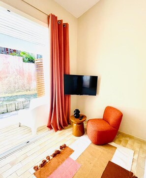 A modern living room with a flat-screen TV, a red armchair, a wooden side table, and a large window with curtains. - Studio Colibri - Terrace 2 pers, 3mn from Anse Mitan beach (Les Trois-Îlets)