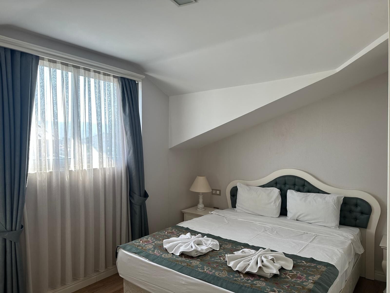 Una habitación de hotel con cama, mesita de noche, lámpara y ventana con cortinas.