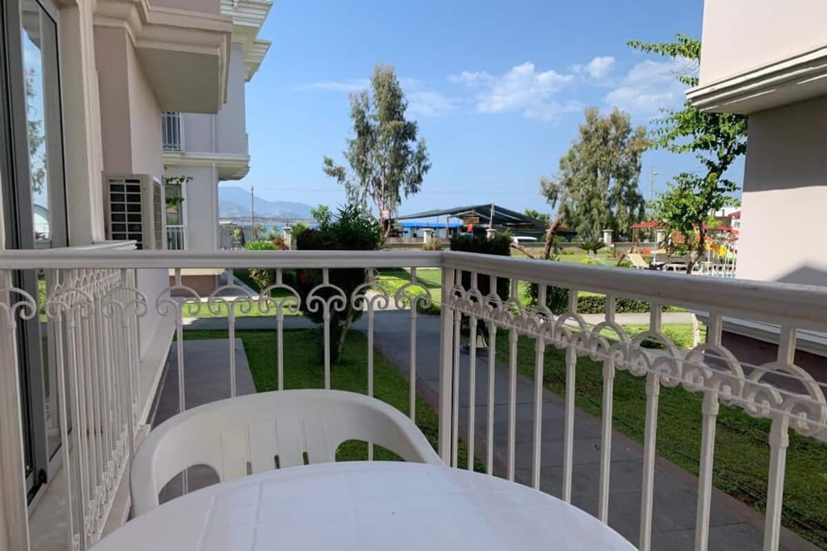 A balcony with a white table and chairs, a metal railing, and a view of a green lawn and buildings in the background.