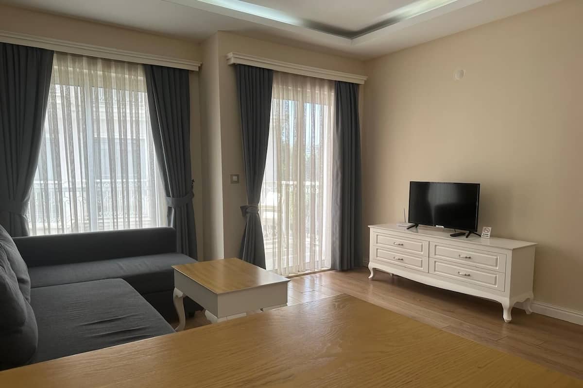 A modern living room with a sofa, a coffee table, a TV on a white dresser, and large windows with curtains.