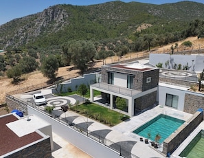 Eine moderne Villa mit Swimmingpool, überdachter Terrasse und Blick auf eine gebirgige Landschaft.