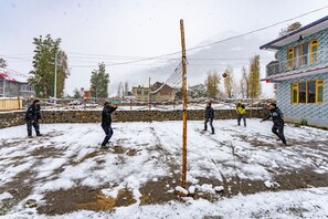 A snowy outdoor area with a volleyball net, stone walls, and buildings in the background. - PACK Sissu (Lahul)