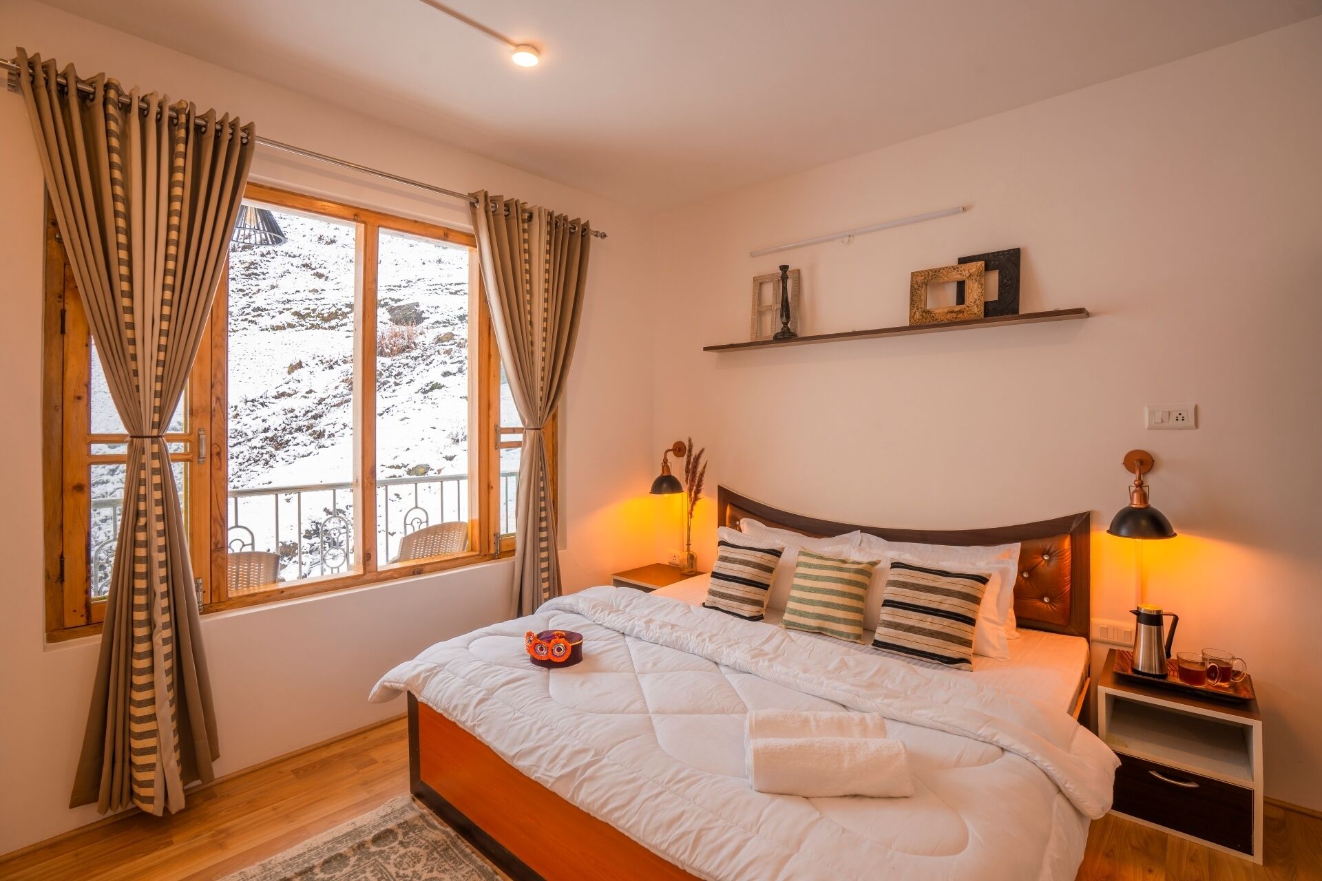Un dormitorio con cama, cortinas, mesita de noche, lámpara y vistas a montañas nevadas a través del ventanal.