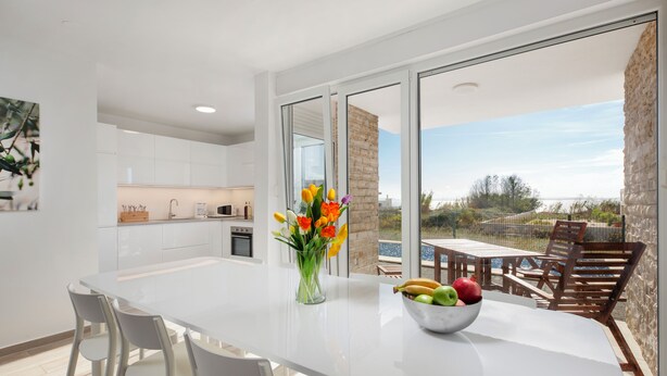 Una cocina moderna con armarios blancos, una mesa de comedor con sillas, un jarrón con flores y un frutero.