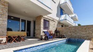 Un área de piscina moderna con pared de piedra, una zona de estar cubierta y una piscina azul cristalina.