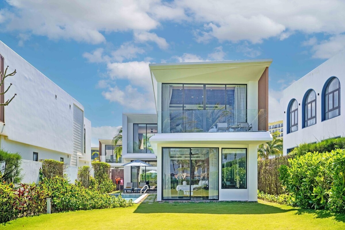 Una casa moderna de dos pisos con un amplio ventanal, piscina y abundante vegetación.