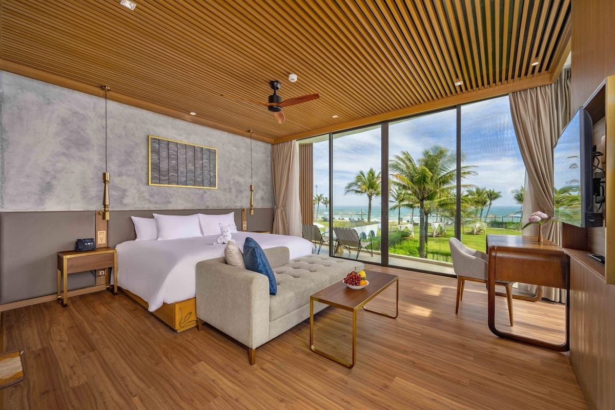 Habitación de hotel moderna con una cama grande, un sofá, una mesa de centro y una televisión. La habitación tiene techo de madera, ventanas amplias con vistas a palmeras y al océano.