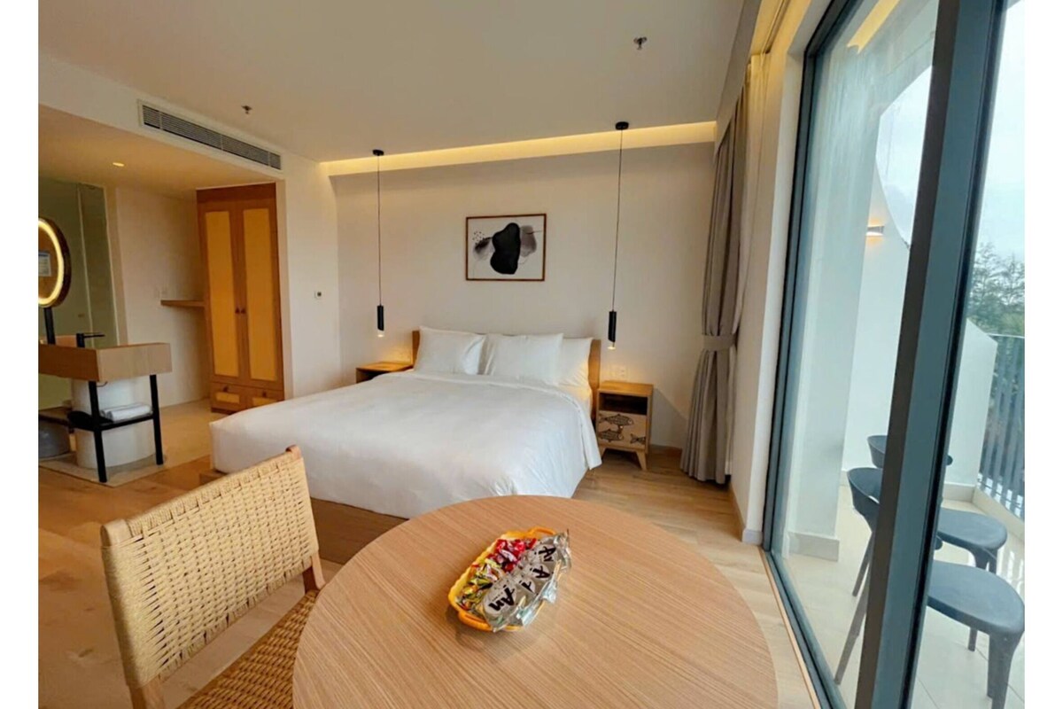 Habitación de hotel con una cama, una mesita redonda con aperitivos, una silla, una mesita de noche, una lámpara, un espejo, una ventana con vistas y una obra de arte colgada en la pared.