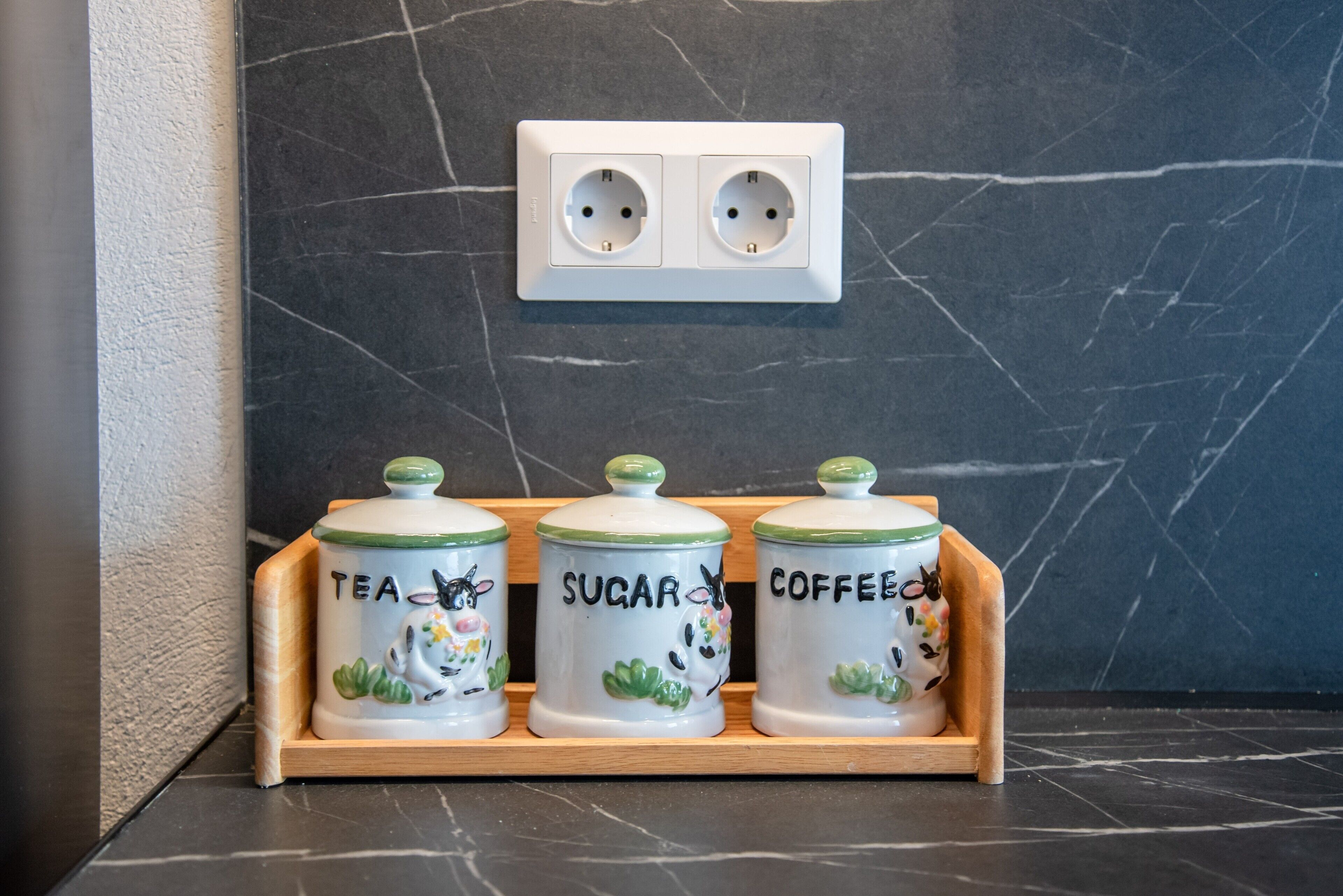 Eine Küchenarbeitsplatte mit drei Keramikkännchen, beschriftet mit 'Tee', 'Zucker' und 'Kaffee', sowie einer Steckdose darüber.