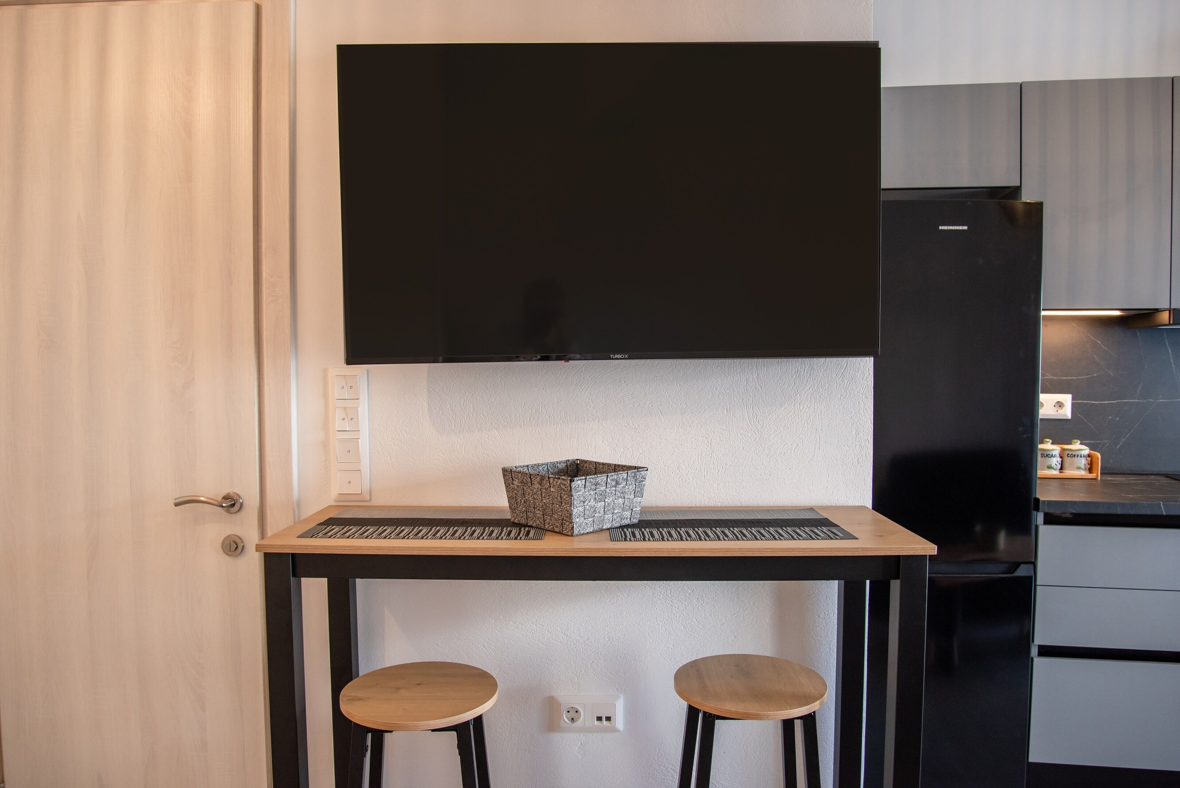 Una cocina compacta con televisor de pantalla plana, un refrigerador negro y una mesa de madera con dos taburetes.