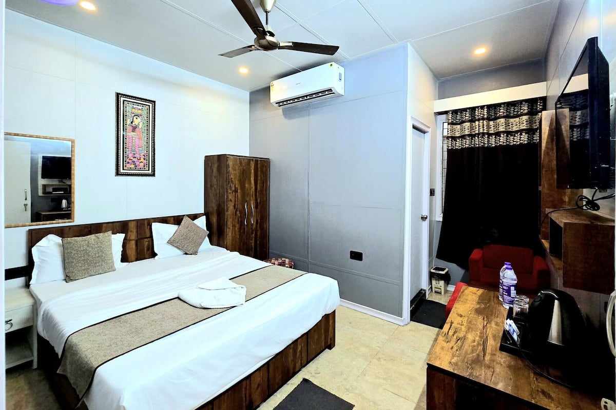 A hotel room with a bed, a wardrobe, a ceiling fan, an air conditioning unit, a television, and a small table with a kettle and a bottle.