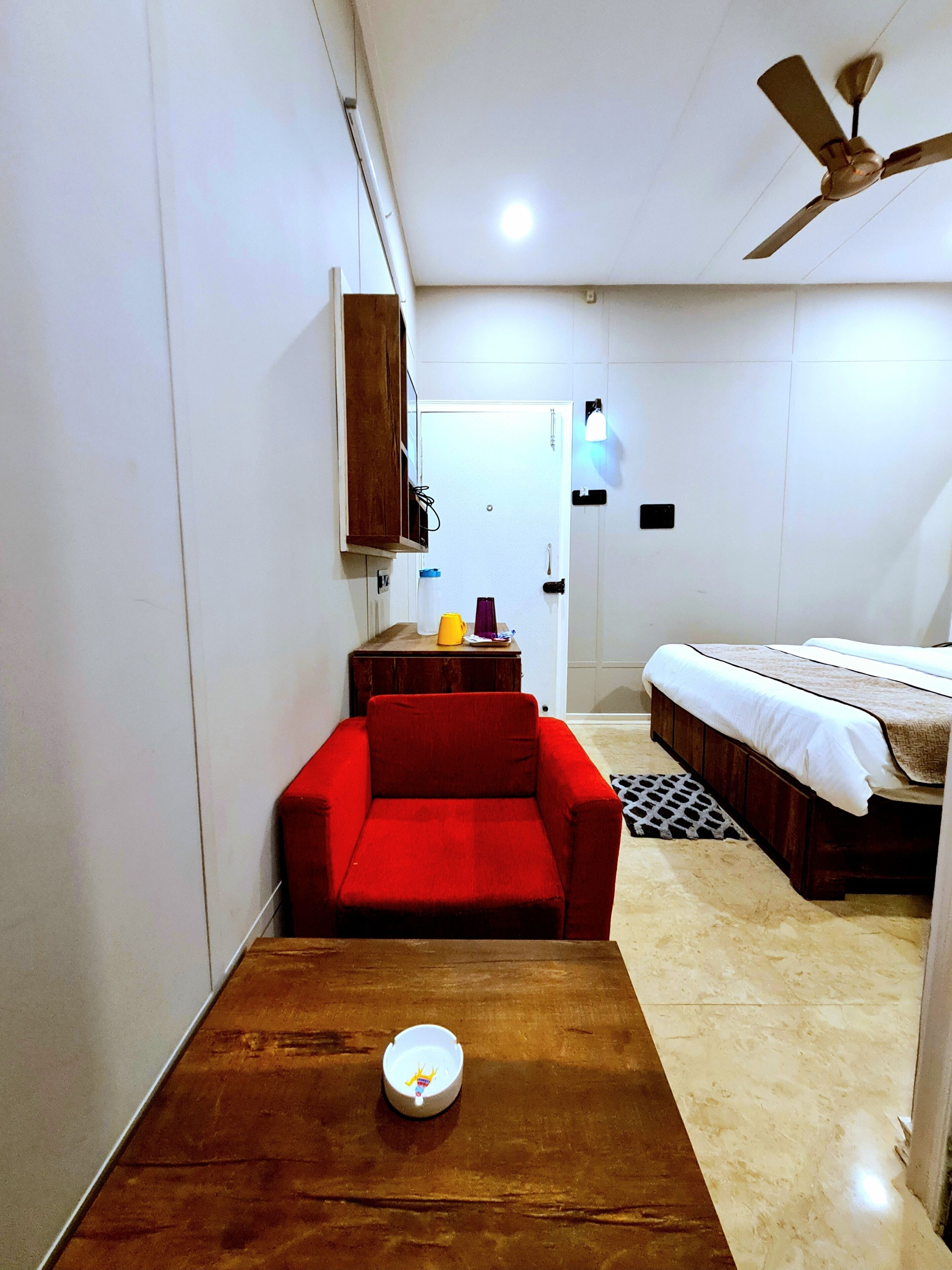Quarto pequeno com cama, sofá vermelho, uma mesinha com uma xícara e ventoinha no teto.