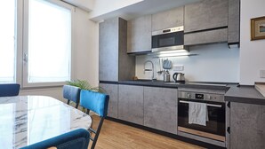 A modern kitchen with grey cabinets, a built-in oven, and a dining area with a marble table and blue chairs. - BRIGHT LUXURY APARTMENT (Milano)