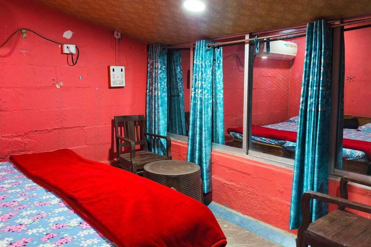 Una habitación con pared roja, una cama con colcha estampada, una silla de madera, una mesita redonda y una ventana con cortinas.