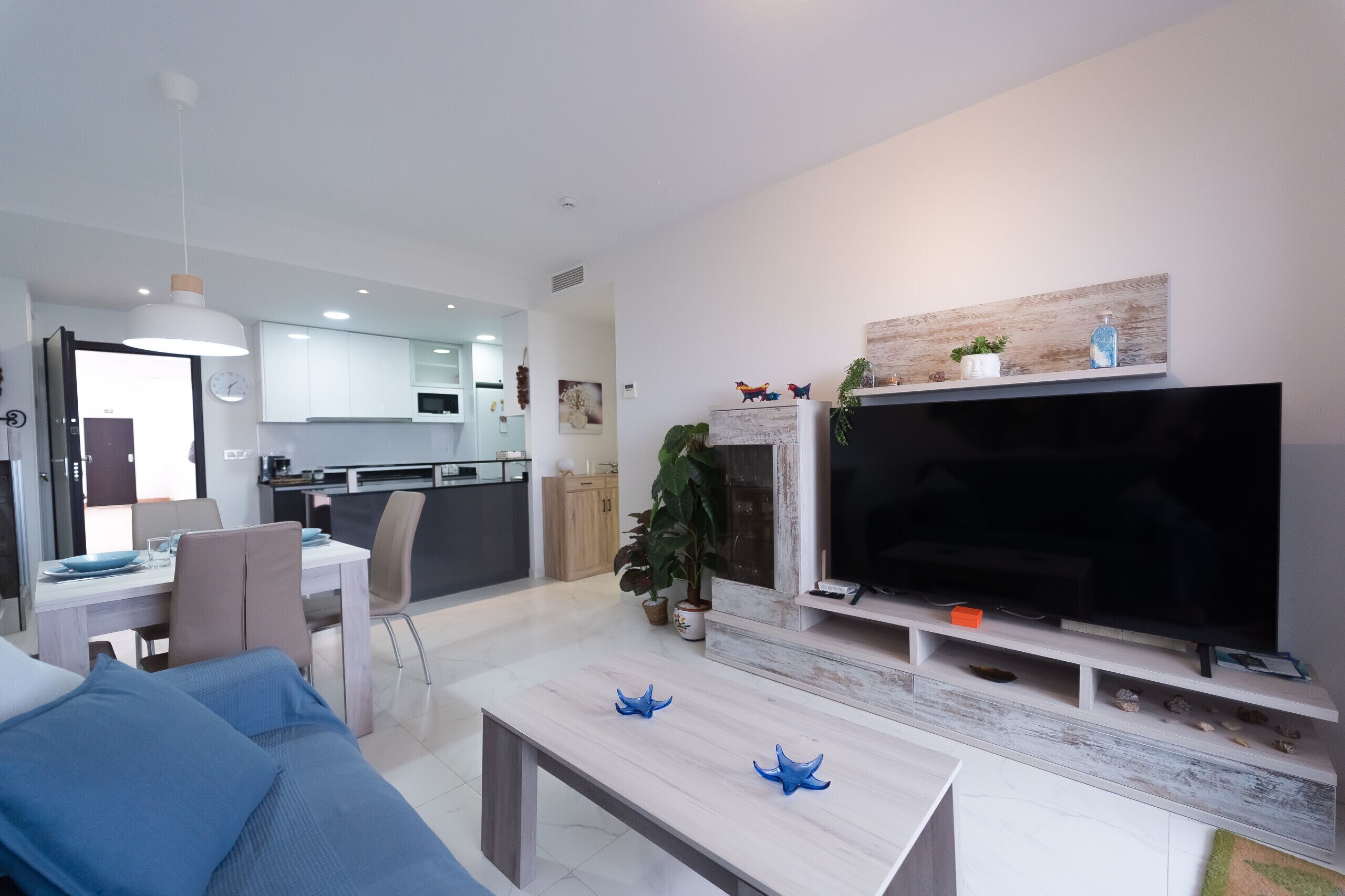 A modern living room with a flat-screen TV, a dining area with a table and chairs, and a kitchen in the background.