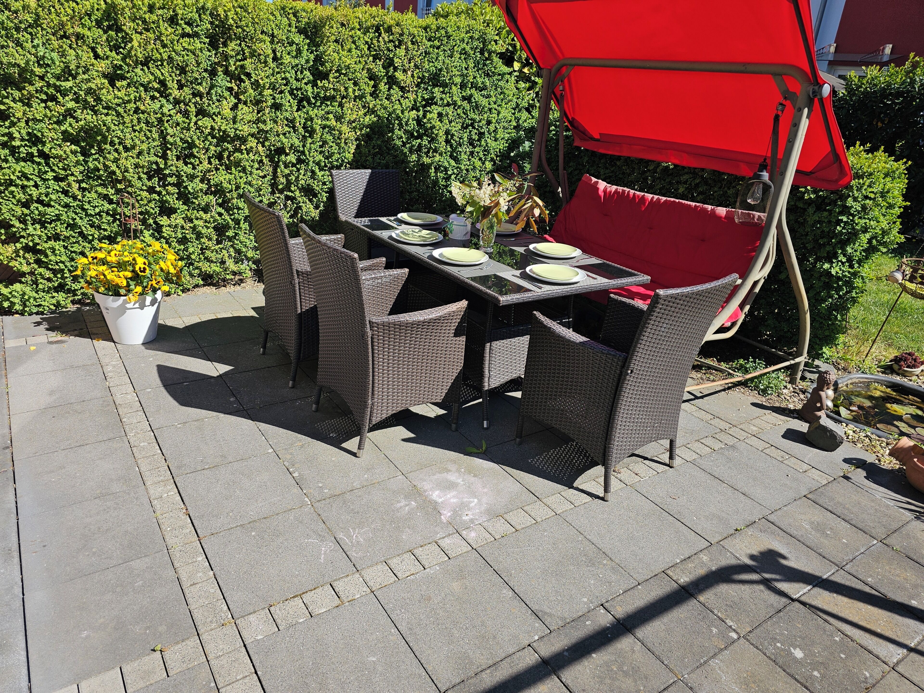 An outdoor patio with a table set for four, surrounded by wicker chairs, a red umbrella, and a potted plant.