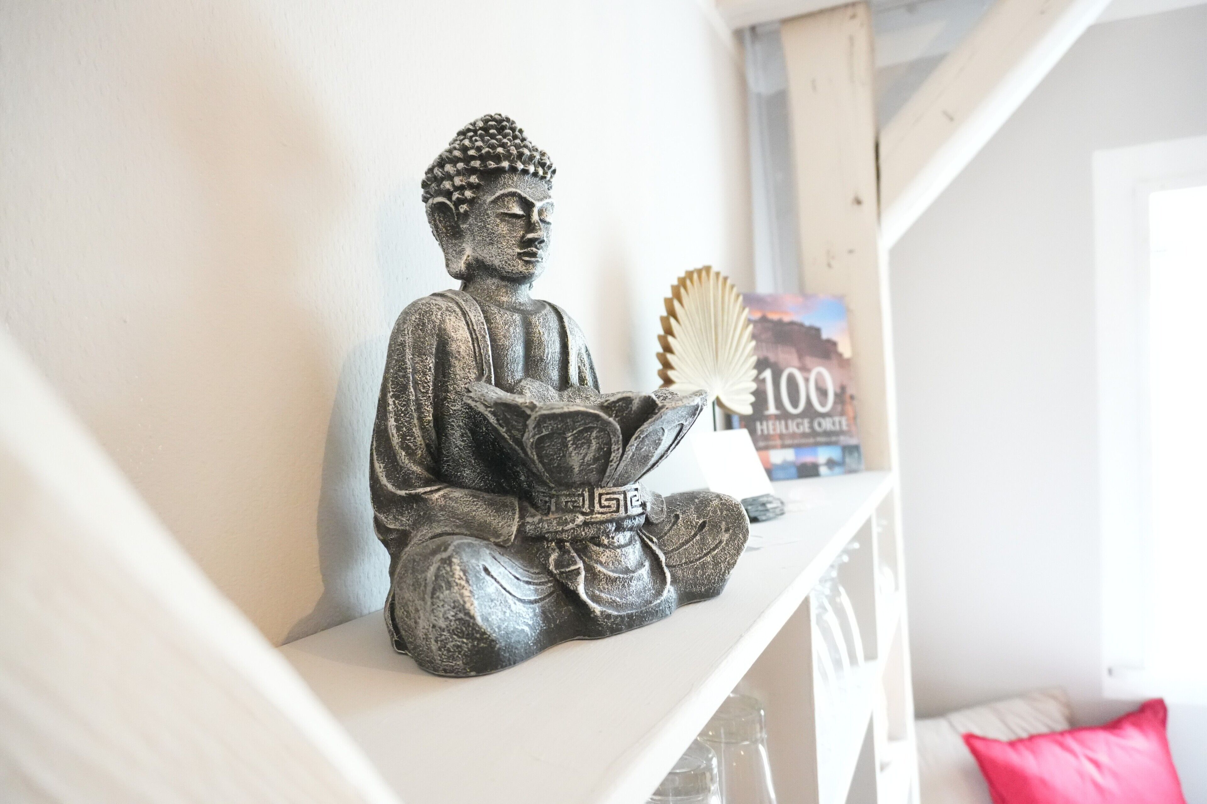 Eine Statue einer sitzenden Figur auf einem Regal, ein Buch mit dem Titel '100 Hellig Ort' und ein rosafarbenes Kissen auf einem weißen Sofa.