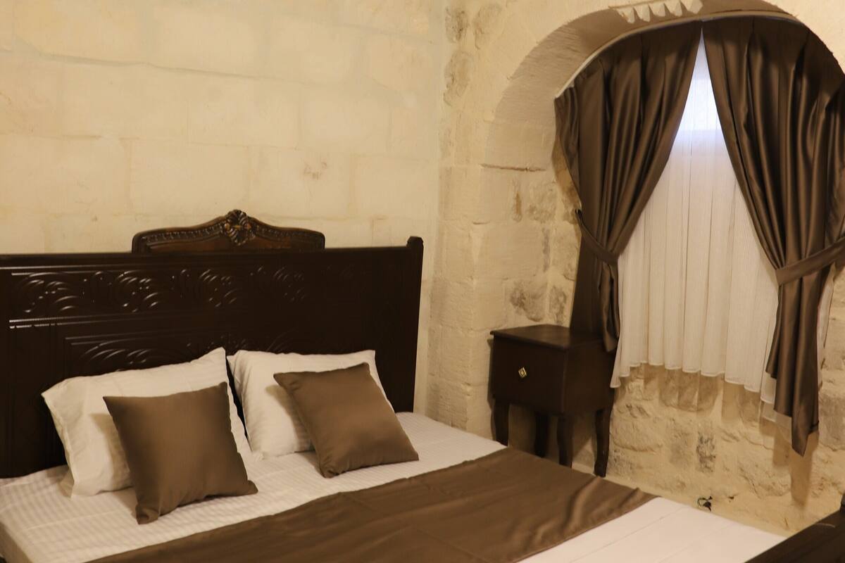 A bedroom with a stone wall, an arched window with curtains, a bed with a headboard, and an air conditioning unit.
