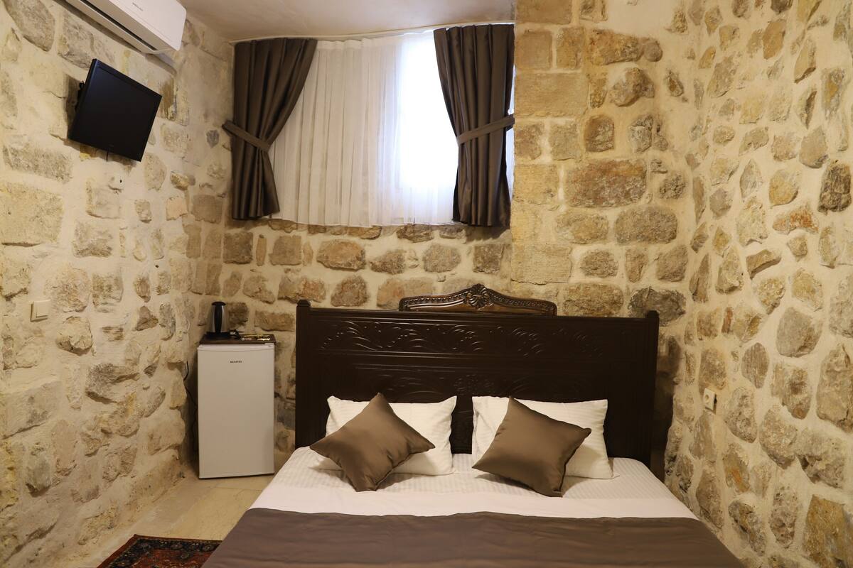 A stone-walled room with a bed, a small white appliance, and a mounted television.