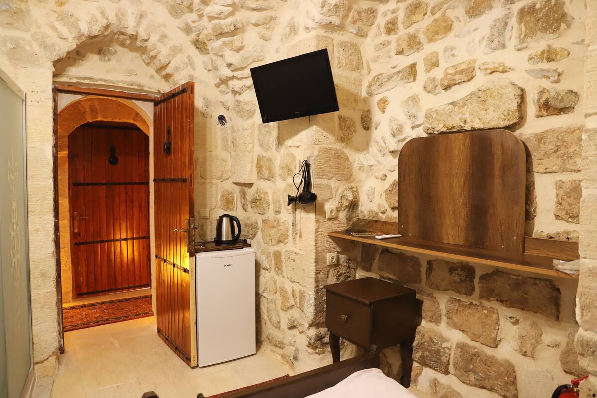 A stone-walled room with a wooden door, a white refrigerator, a flat-screen TV mounted on the wall, and a small wooden cabinet.