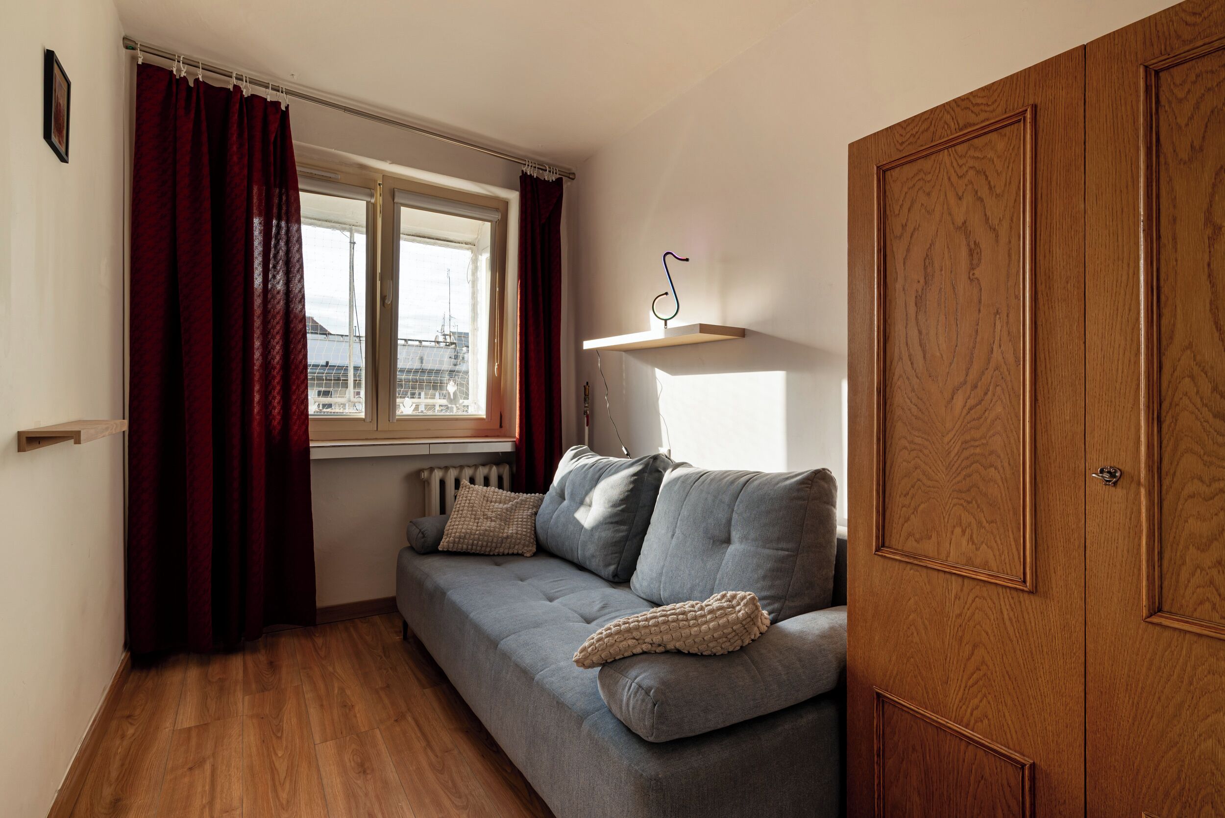Una habitación con un sofá gris, un armario de madera, una ventana con cortinas y un radiador.