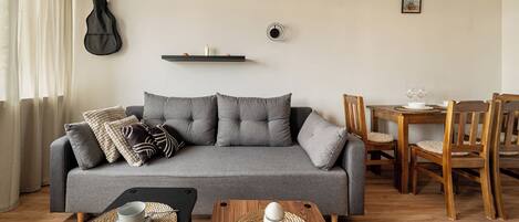 Una sala de estar con un sofá gris, una mesa de comedor con sillas, una guitarra y un estante pequeño con adornos.