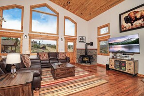 A living room with a fireplace, large windows, a sofa, and a television. - New! Hollow Pine Lodge | Hot Tub + Updated Mountain Retreat Near RMNP (Grand Lake)