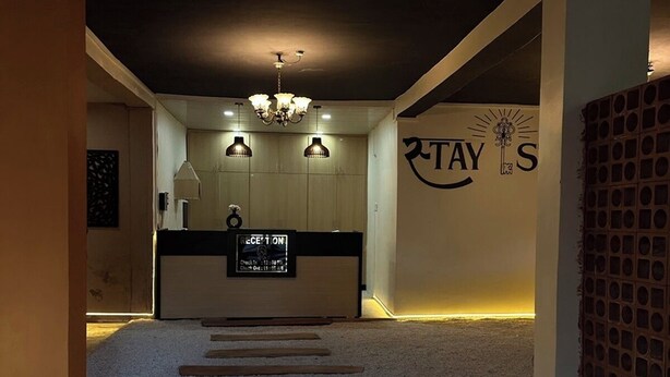 A reception area with a reception desk, a chandelier, a wall with the logo 'STAY'S', and a carpeted floor with wooden planks. - Stay Stories (Chopra)