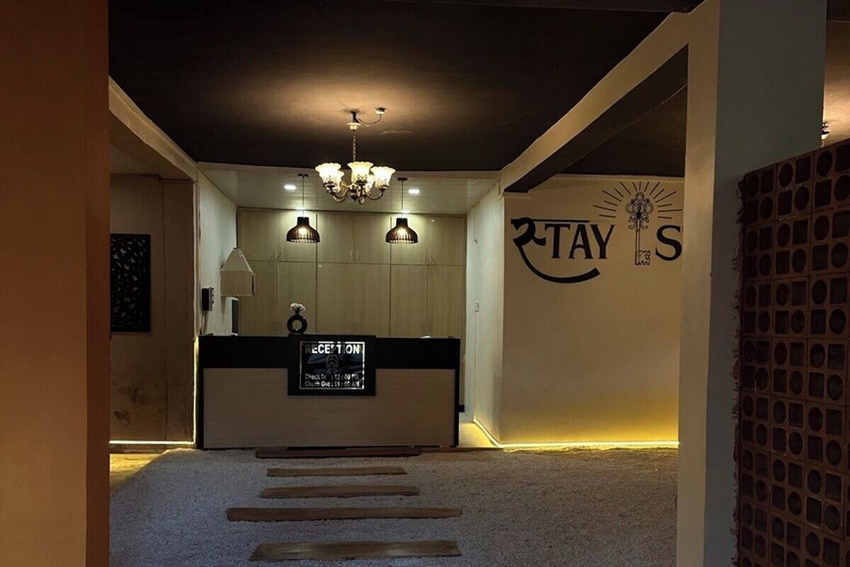 Uma área de recepção com balcão, um candelabro, uma parede com o logotipo 'STAY'S' e piso revestido de carpete com ripas de madeira.