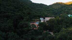 A hillside view of a multi-story building with a mix of residential and commercial structures amidst dense forest. - Stay Stories (Chopra)