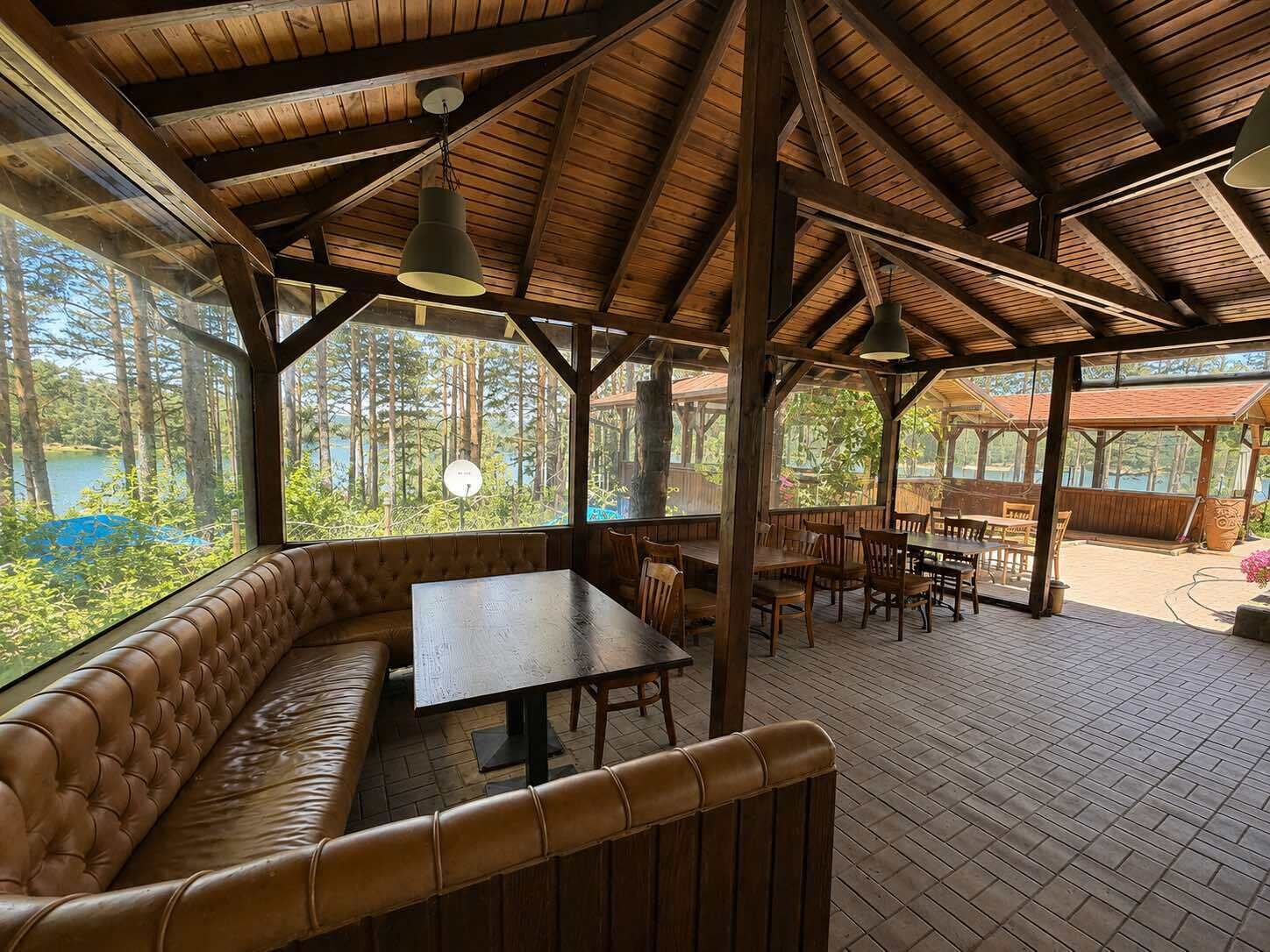 A covered outdoor seating area with a wooden bench and tables, large windows, and a view of a forested area.