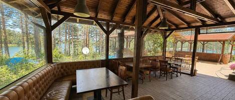 A covered outdoor seating area with a wooden bench and tables, large windows, and a view of a forested area.
