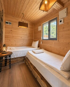 A wooden cabin with two beds, a table with a lantern, and a window with a view of greenery. - Club Paradise Bay Iskar Lake (Sofia)