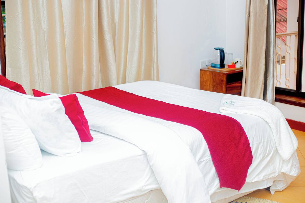 A double bed with white bedding and a red accent, a wooden nightstand with a kettle, and a window with curtains.