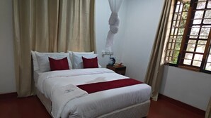 A neatly made bed with white linens and red accents, a bedside table with a lamp and a bottle, and a window with a view of greenery. - kilimanjaro safari lodge (Moshi)