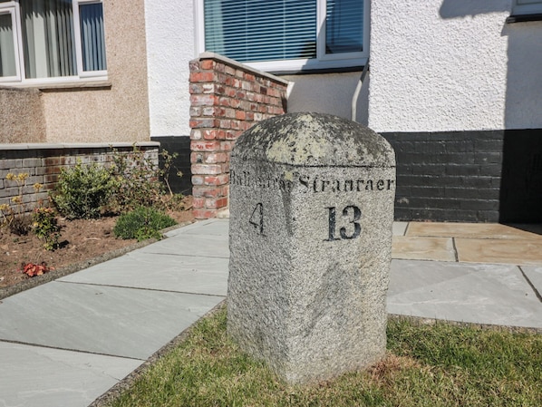 A stone headstone with the inscription 'Balfron Stranaer 4 13' in a landscaped area with a brick wall and a window in the background.