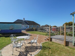 A patio with white metal chairs and a table, a mural of a ship on a wall, and a house with a grey roof in the background.