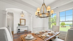 A dining area with a wooden table set for four, white chairs, a chandelier, and a view of a green landscape through large windows. - Kiser Island (Terrell)