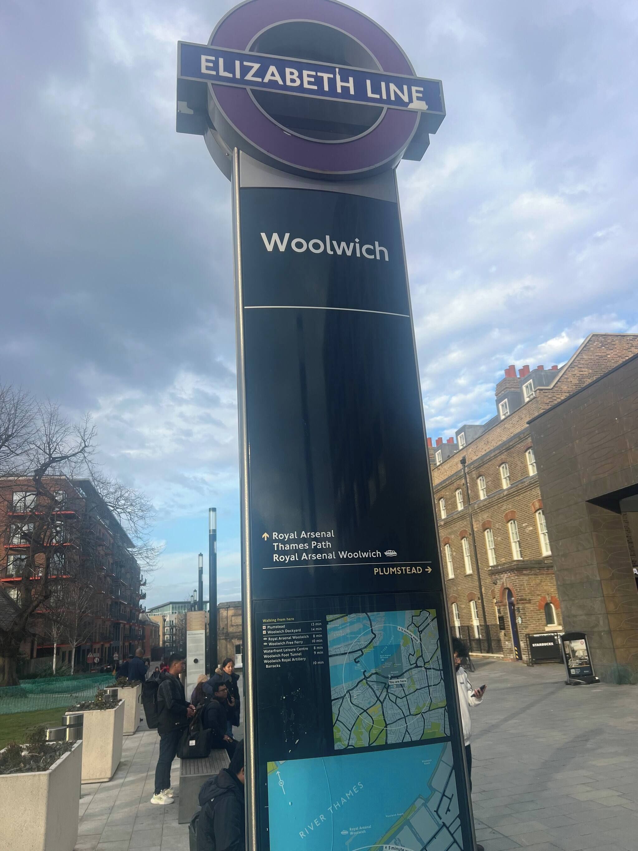 Ein hohes Schild mit der Aufschrift 'ELIZABETH LINE' und 'Woolwich', das einen Bahnhof anzeigt.
