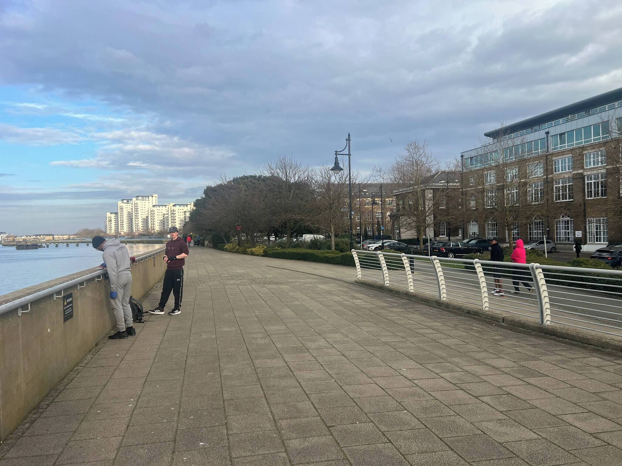Een verharde promenade langs het water met een hekwerk, gebouwen op de achtergrond en een heldere lucht.
