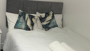 A neatly made bed with white bedding, two decorative pillows, a rolled towel, and a remote control on the bed.