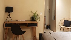 Une chambre avec un bureau en bois, une chaise noire, un lit avec un matelas blanc, une plante en pot et une chaise en osier.