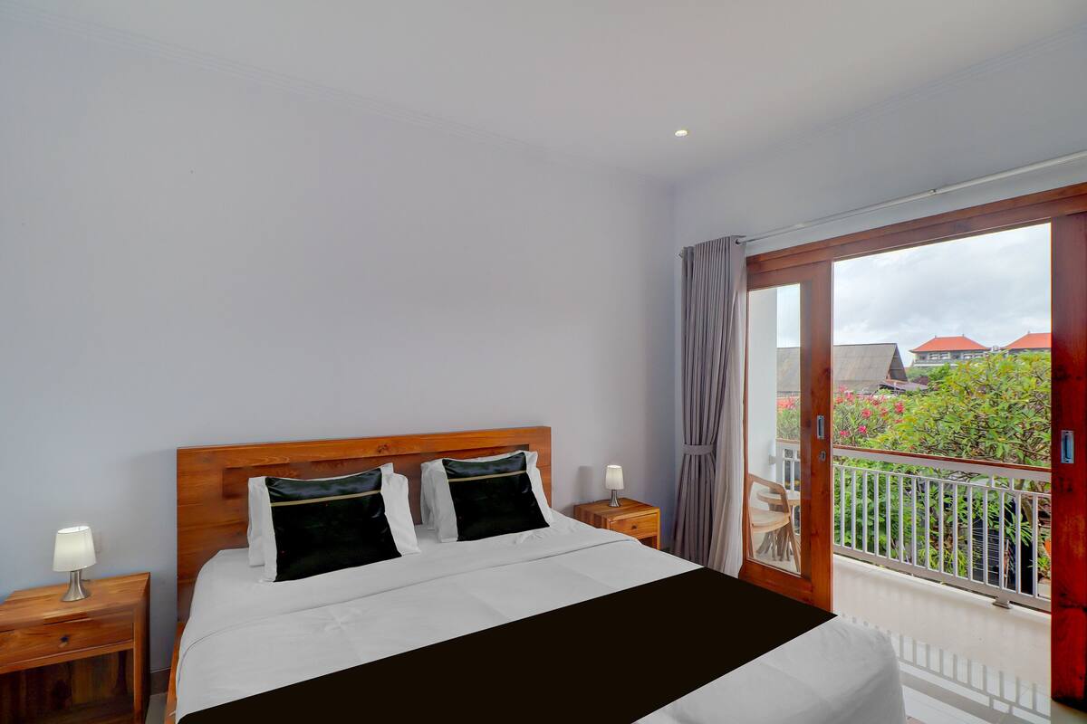 Une chambre d’hôtel comprenant un lit, une tête de lit en bois, deux tables de chevet avec des lampes, un balcon avec une balustrade, et une vue sur la verdure et les bâtiments.