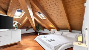 Un dormitorio con techo inclinado, tragaluces, cama, mesita de noche, televisor y cómoda blanca.
