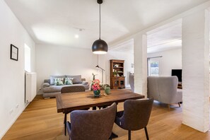 A modern living room with a wooden dining table, two chairs, a sofa, a bookshelf, and a hanging light fixture. - Holiday Home 'Lilla Huset' with Private Terrace and Wi-Fi (Arnsberg)