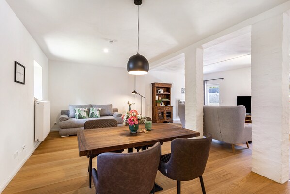 A modern living room with a wooden dining table, two chairs, a sofa, a bookshelf, and a hanging light fixture.