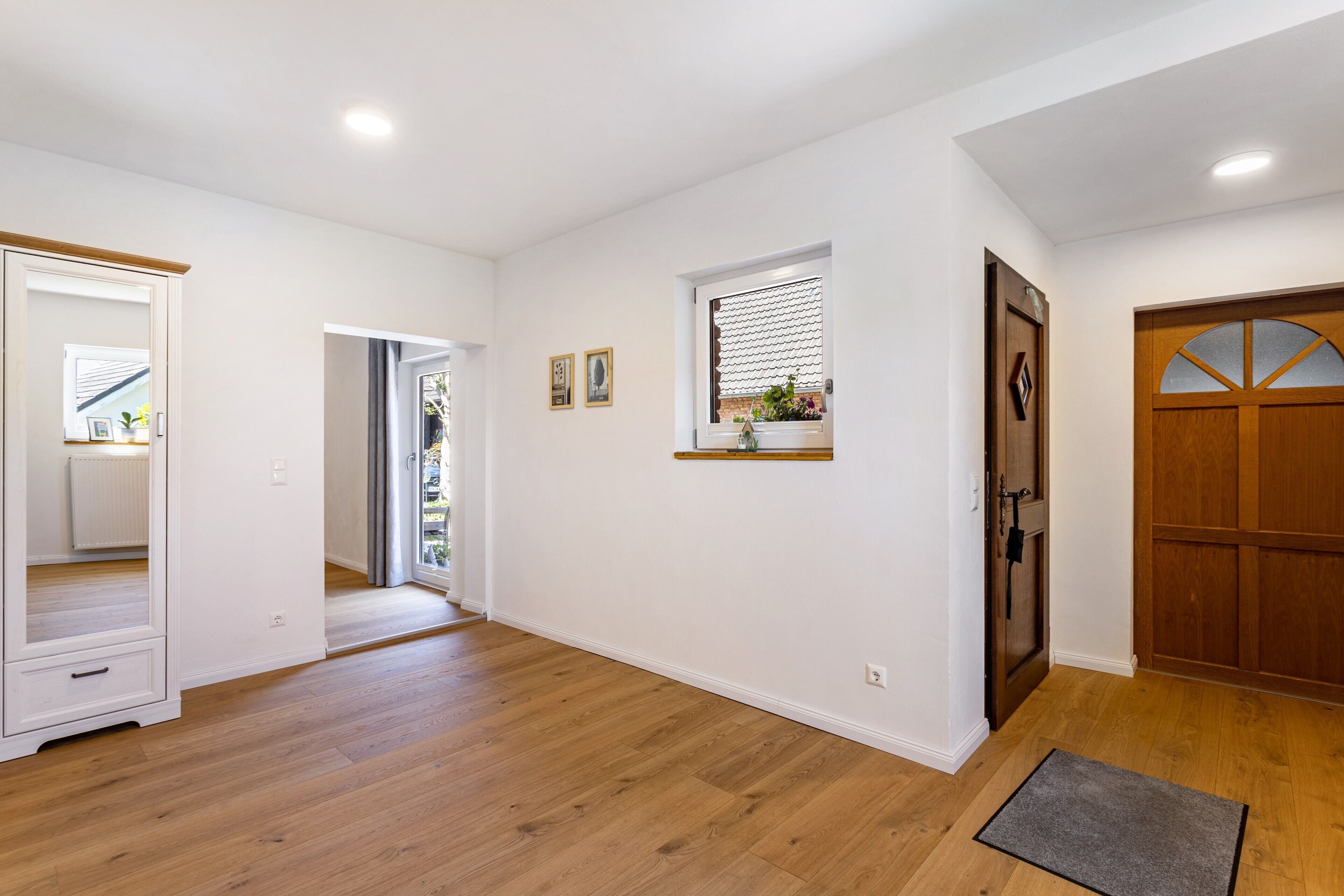 A hallway with wooden flooring, a mirror, a door with a window, and a small shelf with a plant.