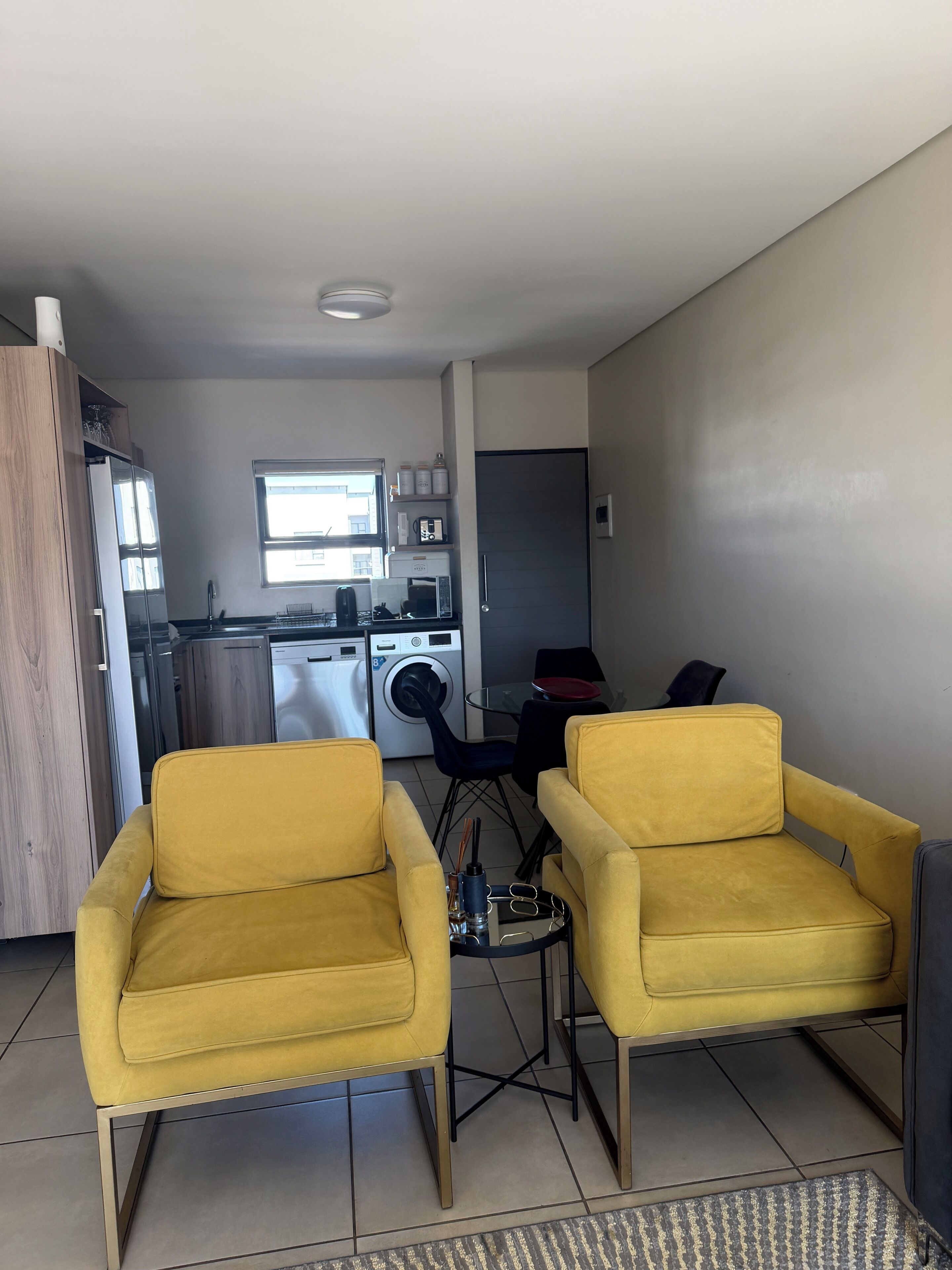 Un espace de vie moderne comprenant une cuisine, deux fauteuils jaunes, une petite table à manger et un lave-linge.