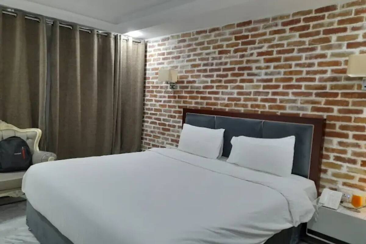 A hotel room with a brick-patterned wall, a bed with white bedding, a nightstand with a phone and a glass, and a couch with a bag.