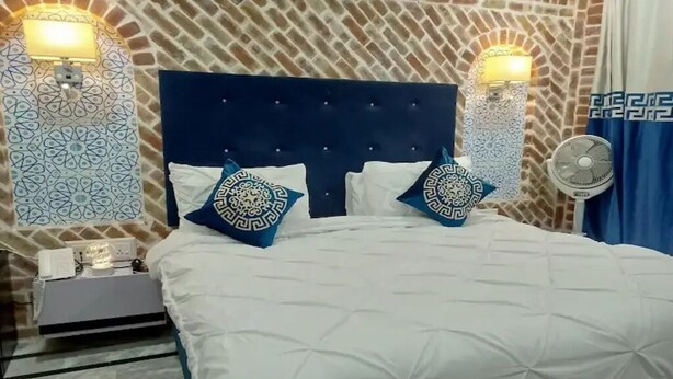 A hotel room with a large bed, two wall-mounted lamps, a nightstand, and a wall-mounted fan. - Hotel Hayatt Sukkur (Sukkur)